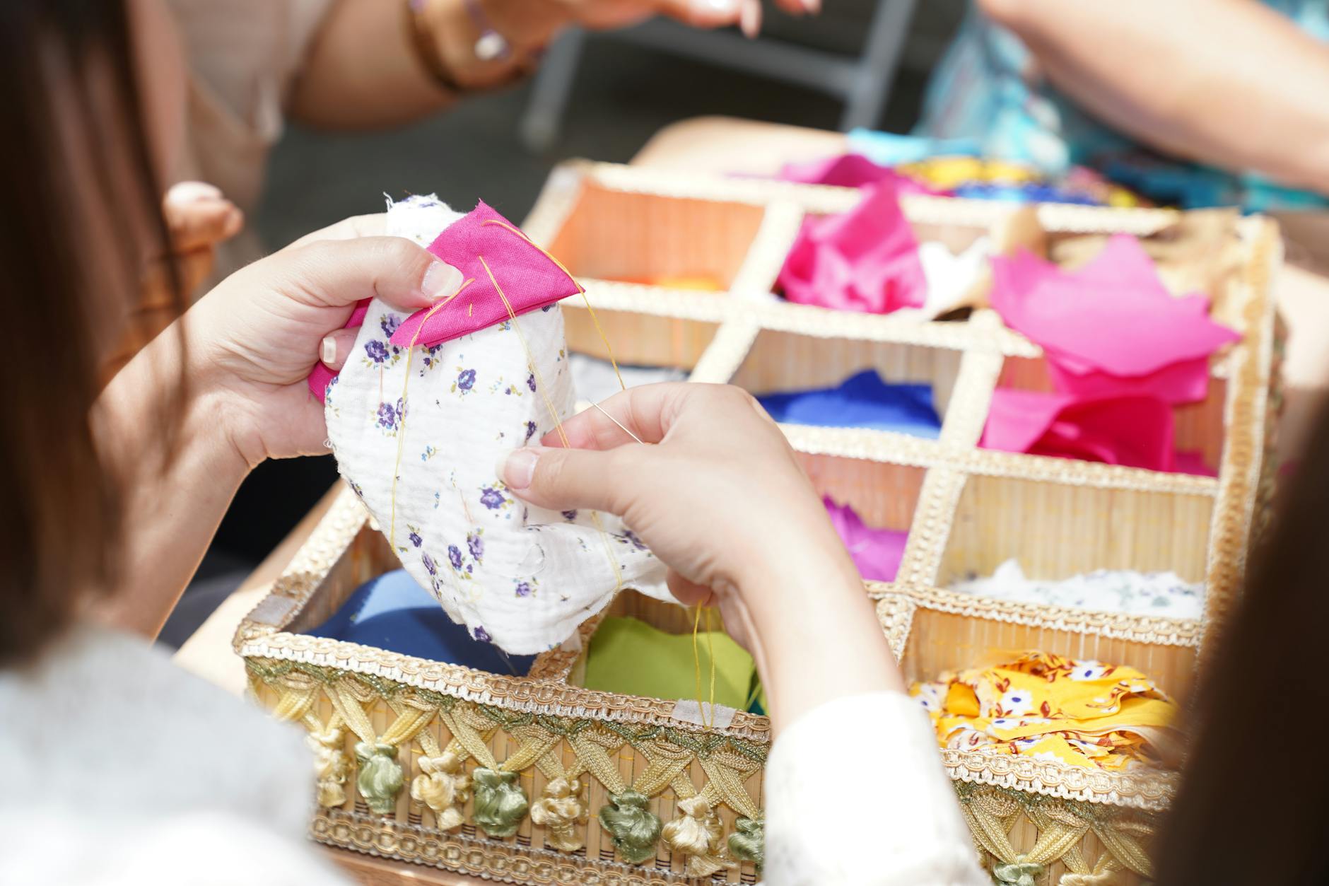 Dłonie szyjące kolorowe skrawki materiału obok wiklinowego pudełka